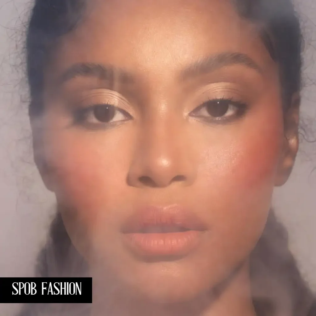 A close-up of a Black woman with a soft-focus 'Cloud Skin' makeup finish. Her skin looks velvet-smooth and airbrushed. She is wearing a soft peach blush and a matching tinted lip balm. The lighting is ethereal and hazy.