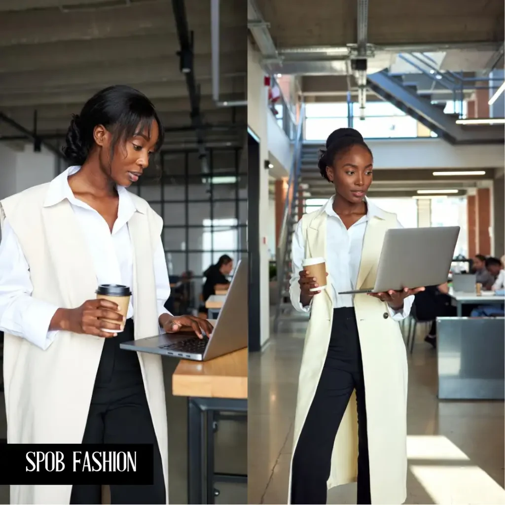 A Black woman in a modern co-working space. She is wearing a cream-colored longline sleeveless vest over a white button-down shirt and black slim-fit trousers. She is holding a coffee cup and looking at her laptop screen.