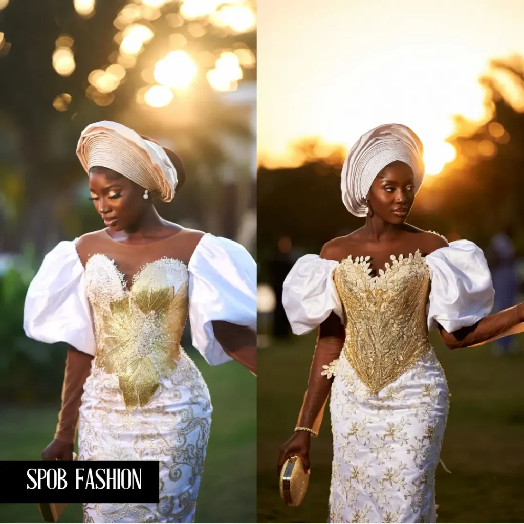 A Black woman at a luxury outdoor wedding. She is wearing a white and gold lace gown with dramatic, oversized butterfly sleeves. She has a high-set Gele and is carrying a gold clutch. The sun is setting behind her, creating a golden halo effect.
