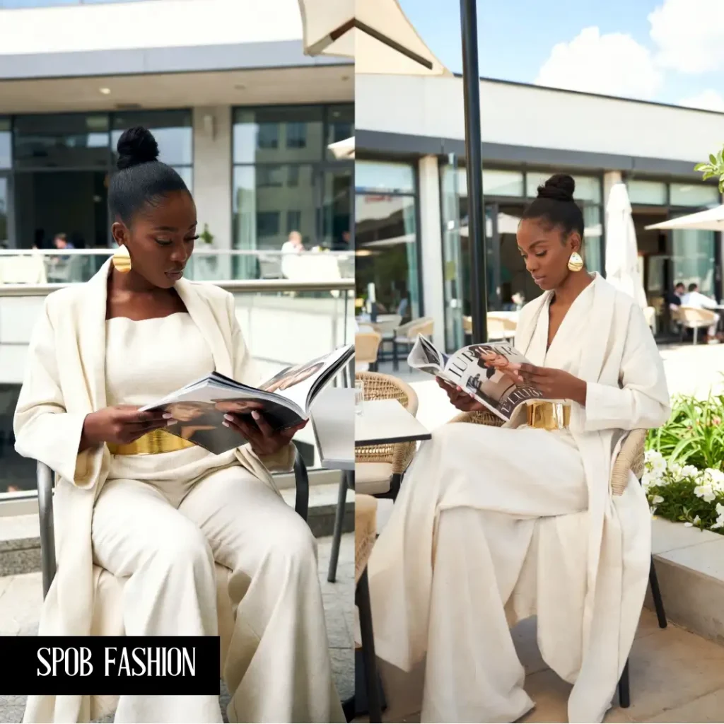 A stylish Black woman at an upscale outdoor cafe. She is wearing a monochromatic cream-colored linen set: wide-leg pants and a long duster coat. She has a bold gold belt and gold hoop earrings. She is reading a luxury fashion magazine. Bright, airy daylight.